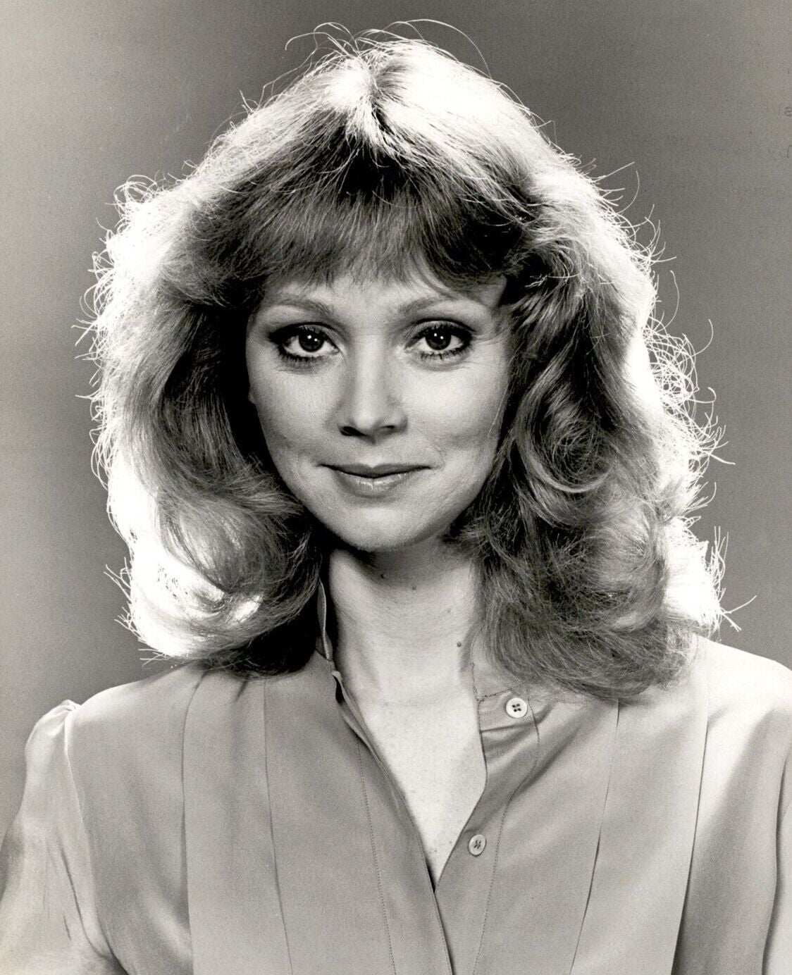 Black and white portrait of a woman with wavy, shoulder-length hair and bangs, wearing a buttoned blouse and smiling softly, looking directly at the camera.