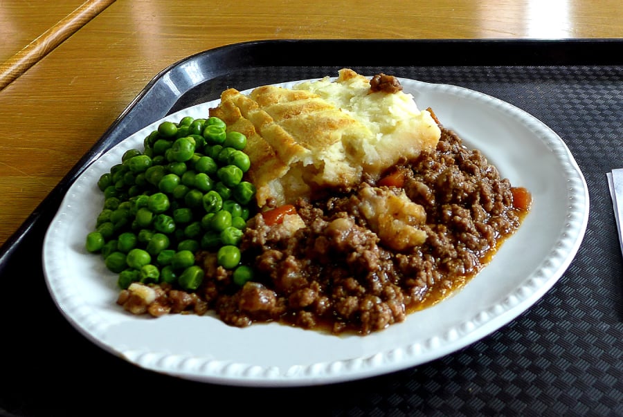 A plate of shepherd's pie with minced meat and mashed potatoes, served with a side of green peas on a white plate placed on a black tray.