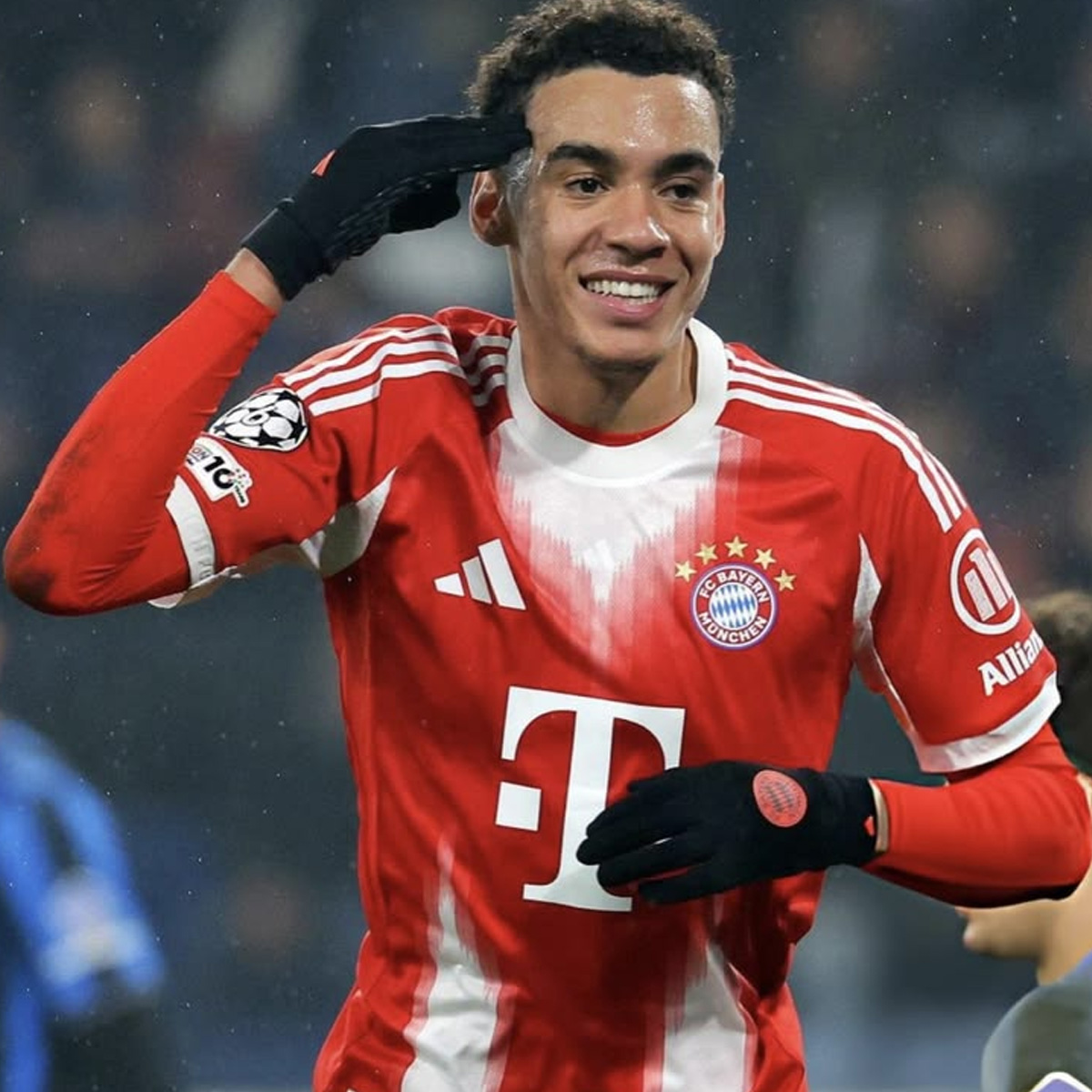 A soccer player wearing a red and white Bayern Munich jersey and black gloves smiles and points to his head with his right hand during a match.