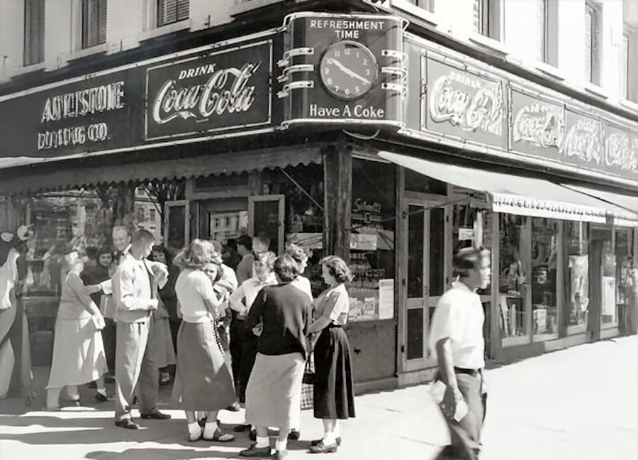 A vintage black-and-white photo shows people gathered outside a corner soda shop with large Coca-Cola signs in the windows and on the awning. Several women in skirts and men in slacks are talking near the entrance.