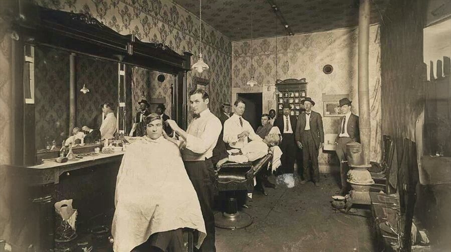 Vintage photograph of a barbershop with barbers cutting customers' hair, while several men in suits and hats stand in the background. The room features patterned wallpaper and antique furnishings.