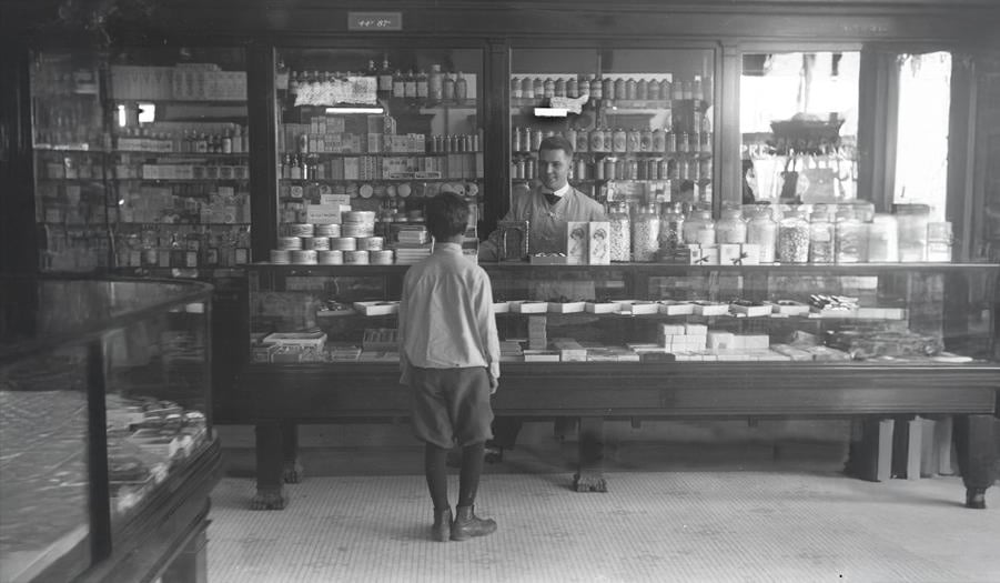 A young boy stands in front of a glass display counter in an old-fashioned shop, facing a man behind the counter surrounded by shelves filled with various goods and jars.