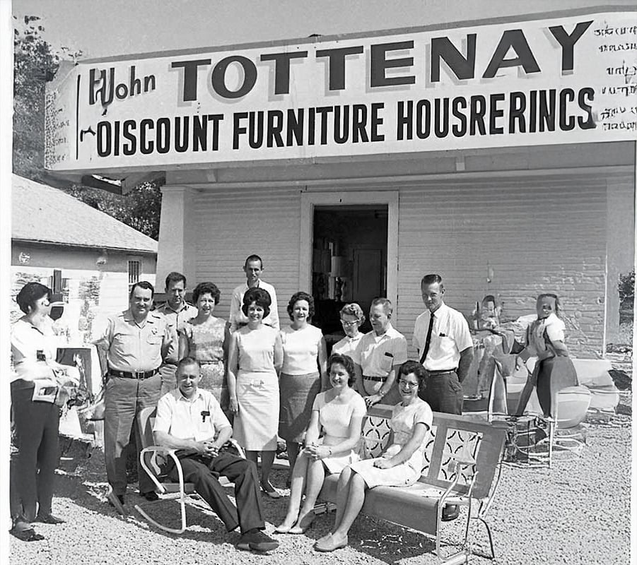 A group of men and women pose outside a store with a sign reading "John Tottenay Discount Furniture Houserenics." Some are seated on patio chairs, others stand, all smiling in front of the storefront.