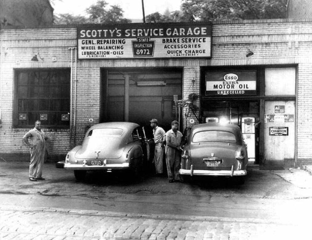 Black-and-white photo of three men standing outside Scotty's Service Garage. Two vintage cars are parked in front of open garage doors beneath signs advertising auto services and motor oil.