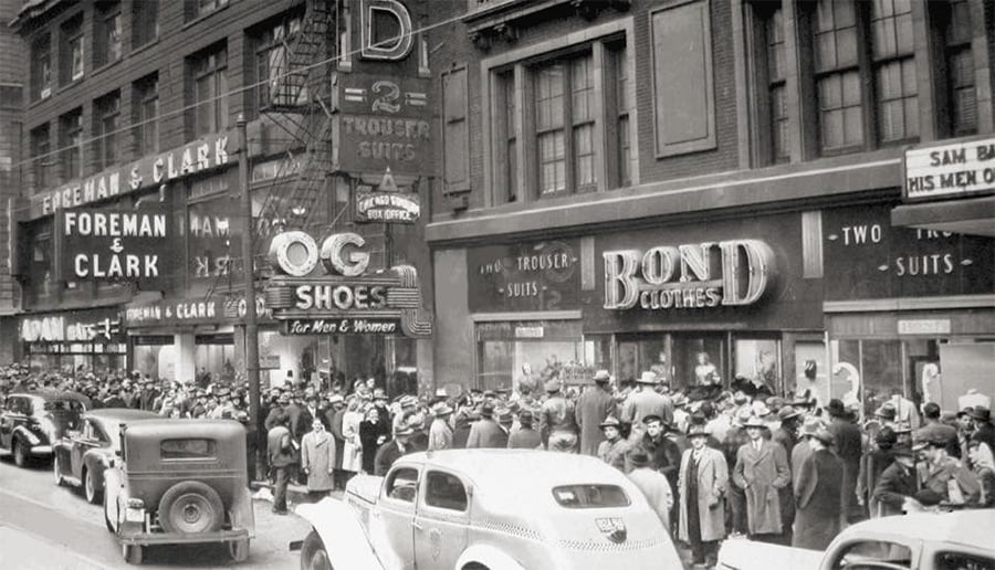 A busy city street in the 1930s, crowded with people in hats and coats. Vintage cars line the road, and storefronts display signs for Foreman & Clark, O.G. Shoes, and Bond Clothes.