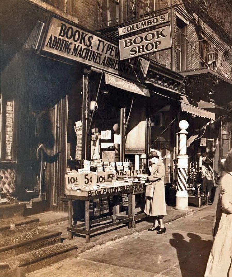A woman browses books displayed on tables outside a vintage bookstore called Columbo’s Book Shop, with dark storefronts, old signs, and a barber pole visible on a city sidewalk.
