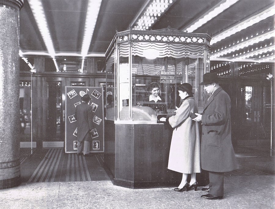 Black-and-white photo of a vintage movie theater entrance. A woman in a ticket booth helps a female customer, while a man waits in line. Another person stands at a door decorated with movie posters.