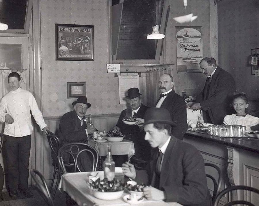 Black-and-white photo of a vintage café with men in suits at tables, a waiter serving food, a chef by the door, and a woman seated at the counter; posters and plates decorate the walls.