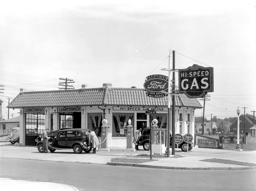Black and white photo of a vintage gas station with two cars at the pumps; attendants assist customers beneath a tiled roof. Signs read “Hi-Speed Gas” and “L.A. Steinert, Ford Specialist, All Makes.”