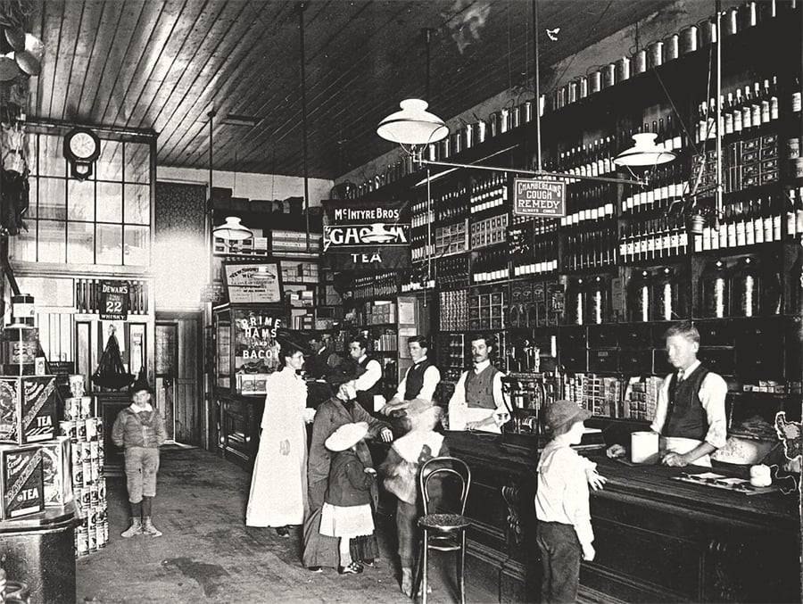 Black-and-white photo of an old-fashioned general store with shelves full of goods, several men behind the counter, and women and children shopping. High ceilings, hanging lights, and vintage signs add to the historic atmosphere.