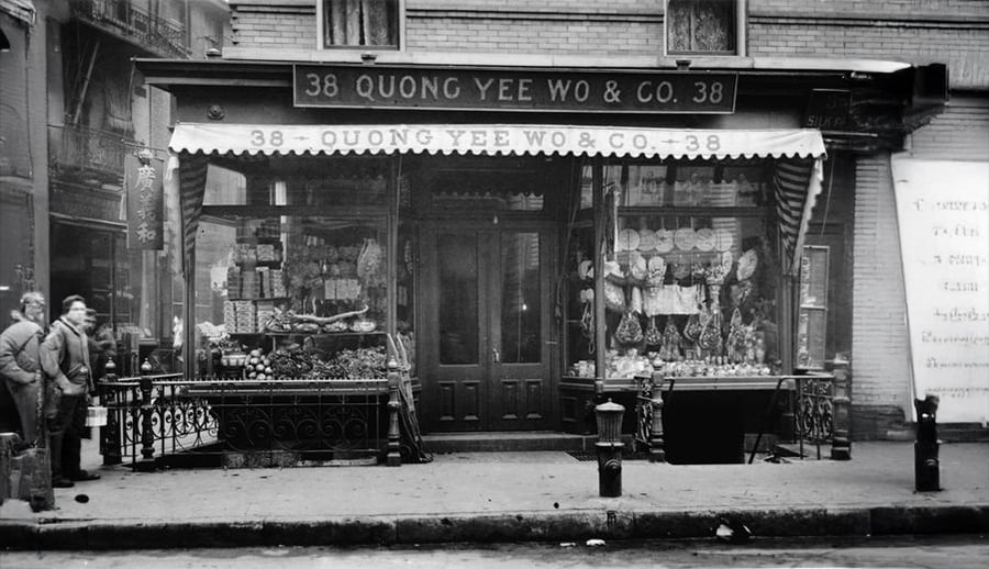 Black-and-white photo of the storefront "Quong Yee Wo & Co. 38" with goods displayed in the window and two people standing outside on the sidewalk. The shop has striped awnings and Chinese characters on the sign.