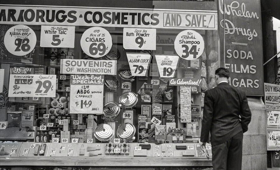 A man in a suit looks at a crowded drugstore window display filled with signs advertising sales on cosmetics, souvenirs, cigars, and toiletries, with prices prominently featured.