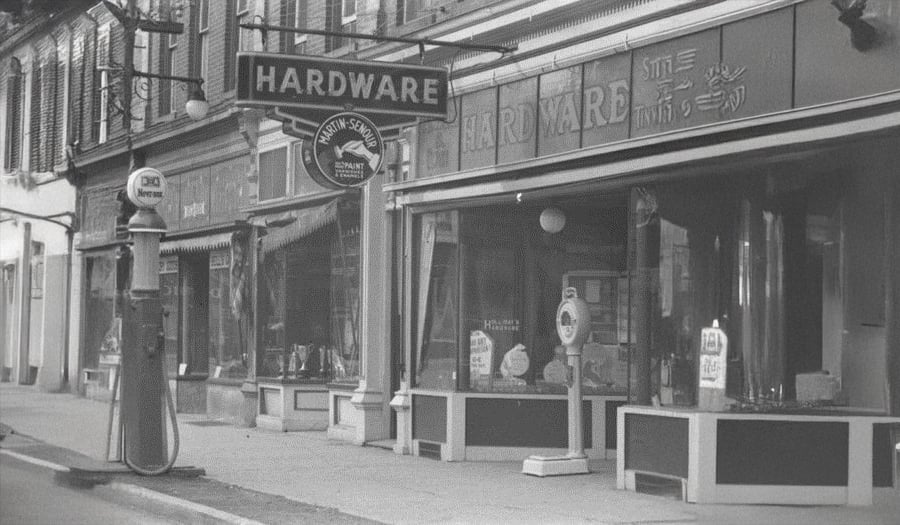 Black and white photo of a vintage hardware store with a large "HARDWARE" sign, storefront windows displaying various items, and an old-fashioned gas pump on the sidewalk in front.