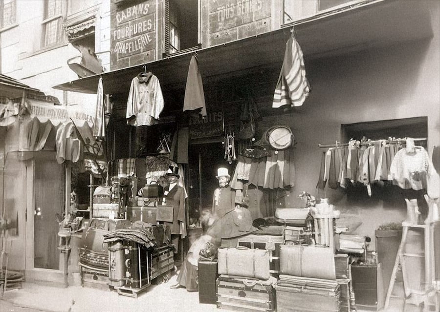 A vintage black-and-white photo of a shop selling trunks, suitcases, and clothing, with various items displayed outside. Three people, two men in uniform and one woman, stand at the entrance. Signs and stacked luggage are visible.
