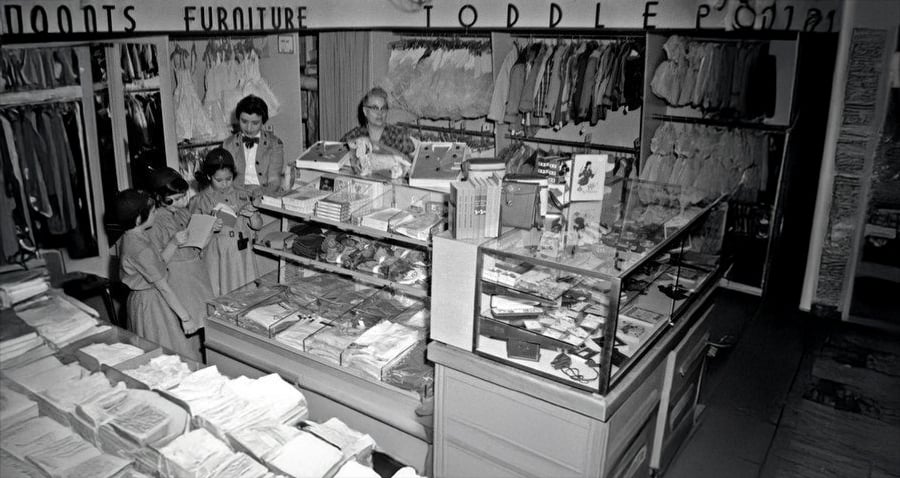 Black-and-white photo of a vintage clothing store with women and children browsing racks and glass display cases filled with folded clothes and small items. Shelves and hanging clothes are visible in the background.