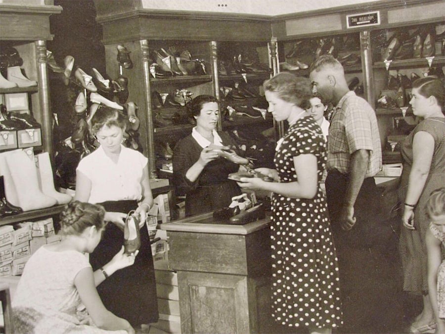 A vintage shoe store scene with women and a man shopping. A clerk stands behind the counter helping customers, while others examine shoes. Shelves behind are filled with various shoes and boxes. The image appears to be from the mid-20th century.