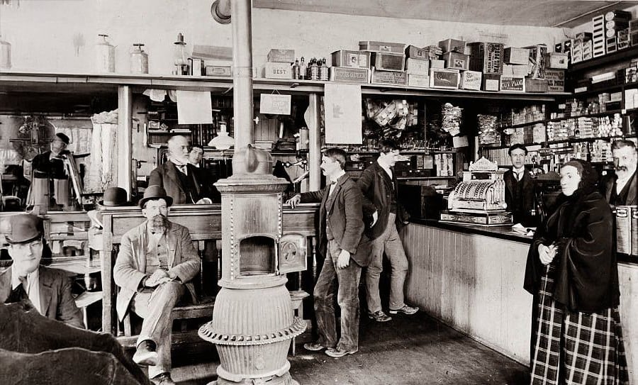 A black and white photo of people inside an old-fashioned general store, with men standing and sitting near a central stove, shelves filled with goods, and a large cash register on the counter.