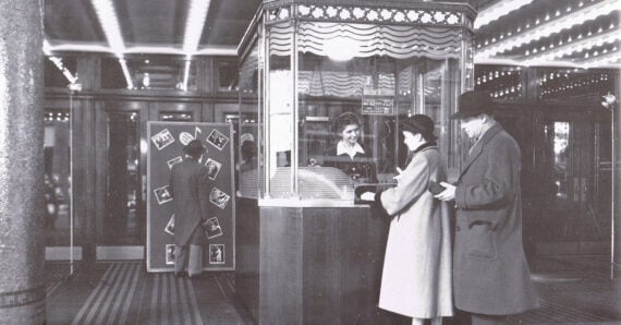 Black and white photo of two people buying tickets at a vintage movie theater box office, with a ticket clerk behind glass and a person looking at movie posters in the background.