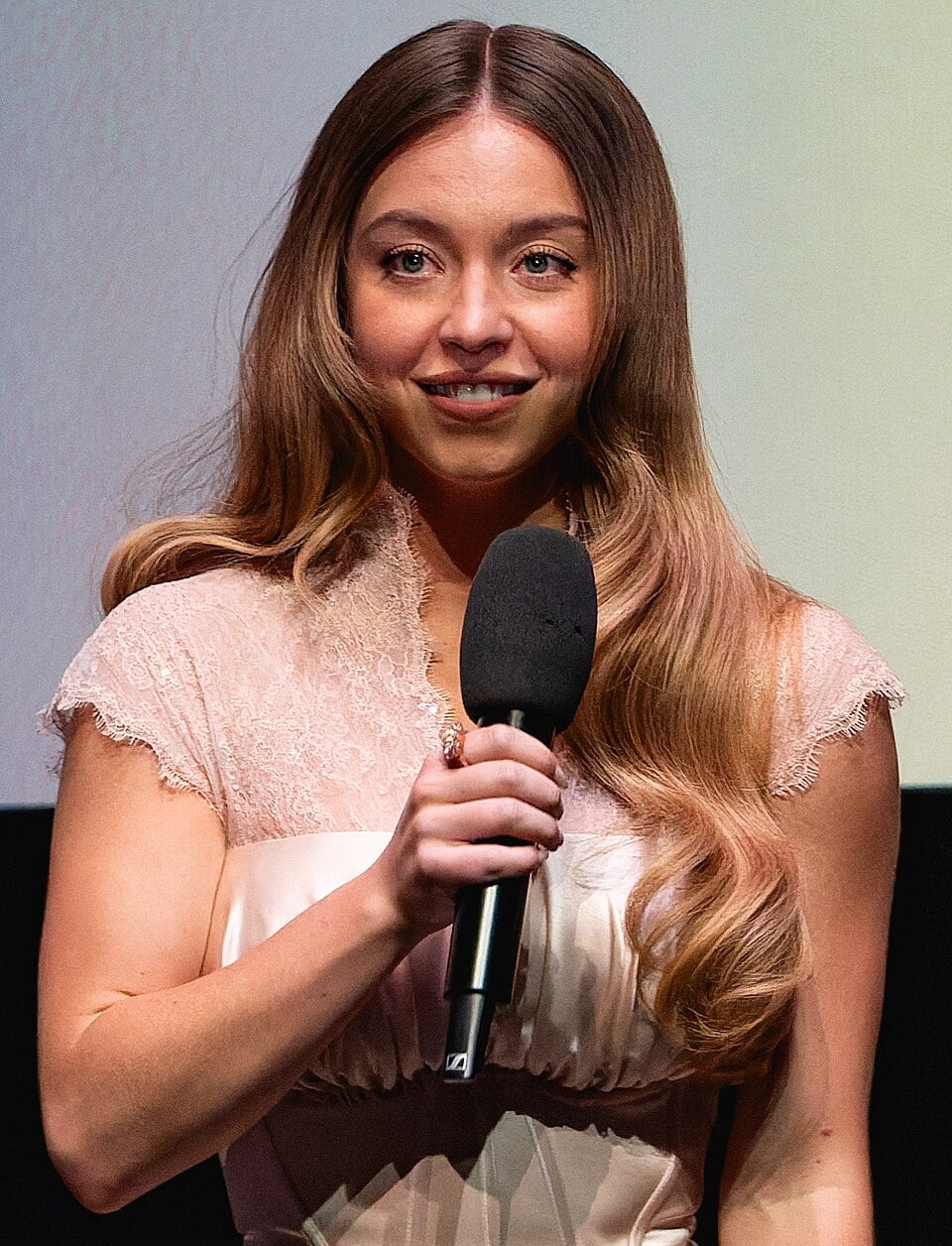 A woman with long, wavy light brown hair wearing a pale pink dress is holding a microphone and smiling while standing indoors against a light and dark background.