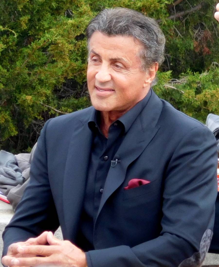 A man with short dark hair and a dark suit with a red pocket square sits outdoors, smiling. Green foliage is visible in the background.