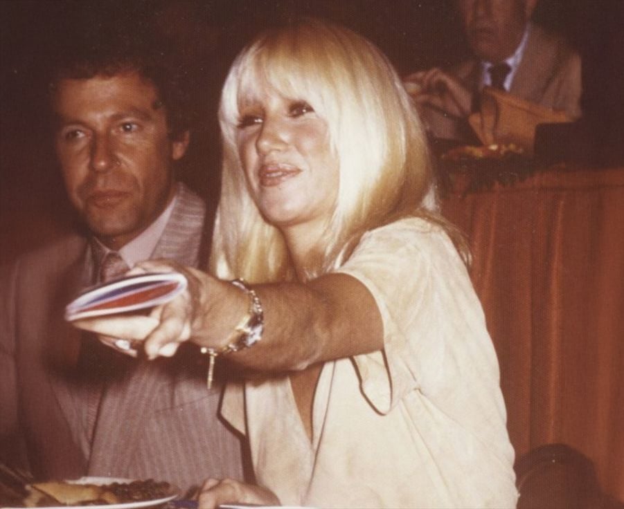A blonde woman sitting at a table, smiling and reaching forward while holding a round object, with a man in a suit seated beside her and another man in the background.