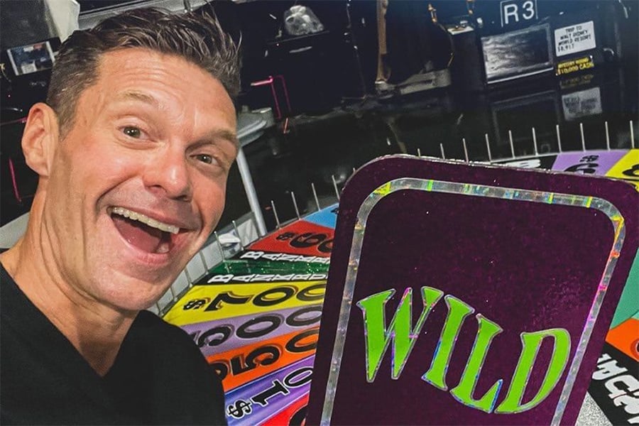 A smiling man takes a selfie next to a colorful game wheel, with a large card in the foreground displaying the word "WILD" in bold letters.