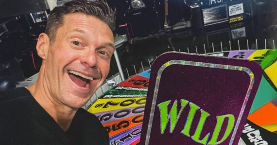 A smiling man takes a selfie in front of a colorful game wheel, holding a large sign that reads "WILD" in bold letters.
