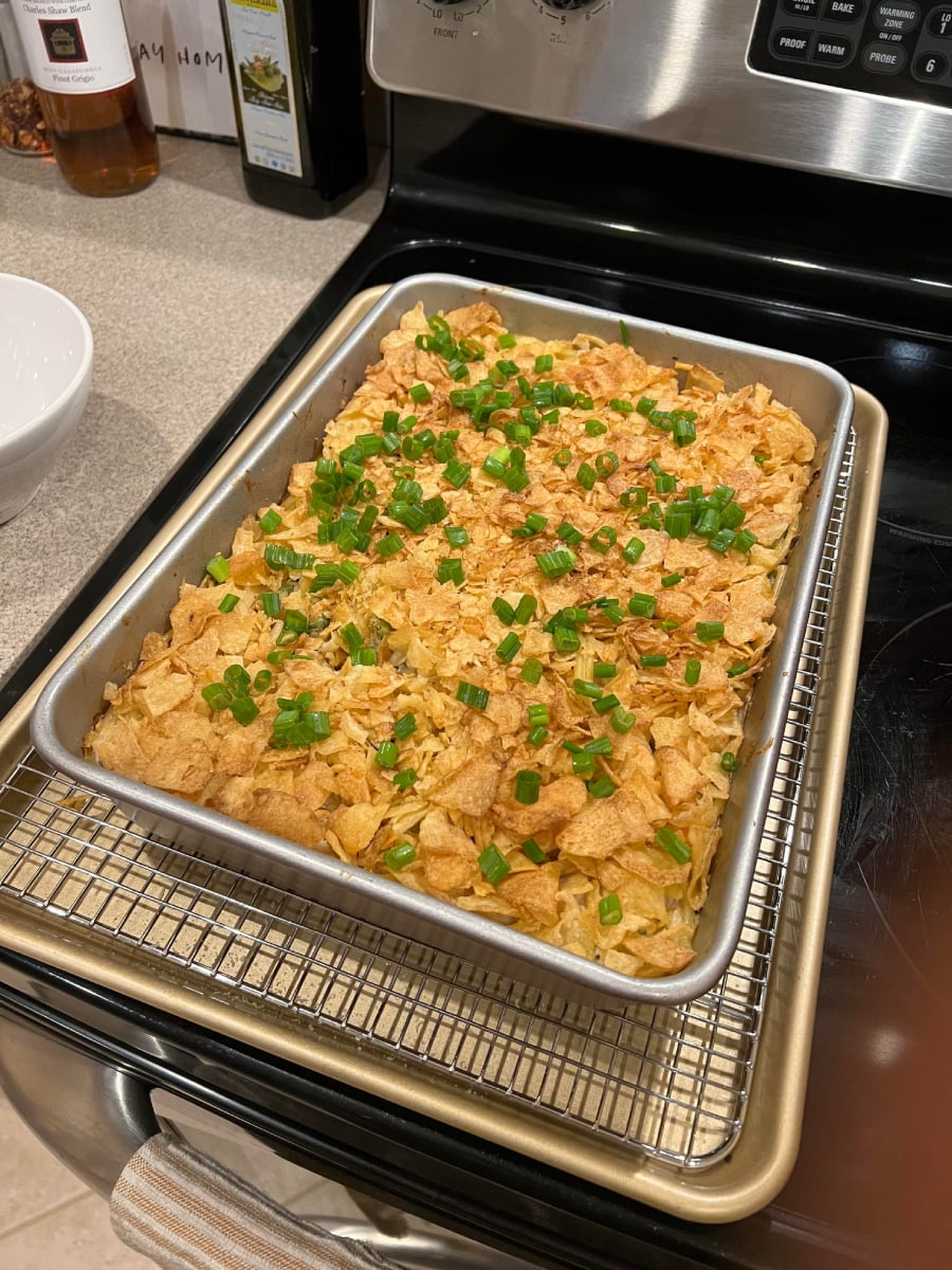 A rectangular baking pan filled with a casserole topped with crushed chips and chopped green onions, resting on a wire rack over a baking sheet on a kitchen stove.