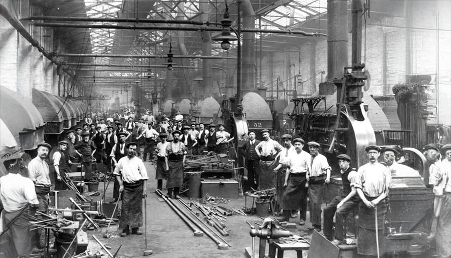Black and white photo of a busy factory with many workers standing among machinery and tools, wearing work clothes and hats, inside a large, high-ceilinged industrial building.