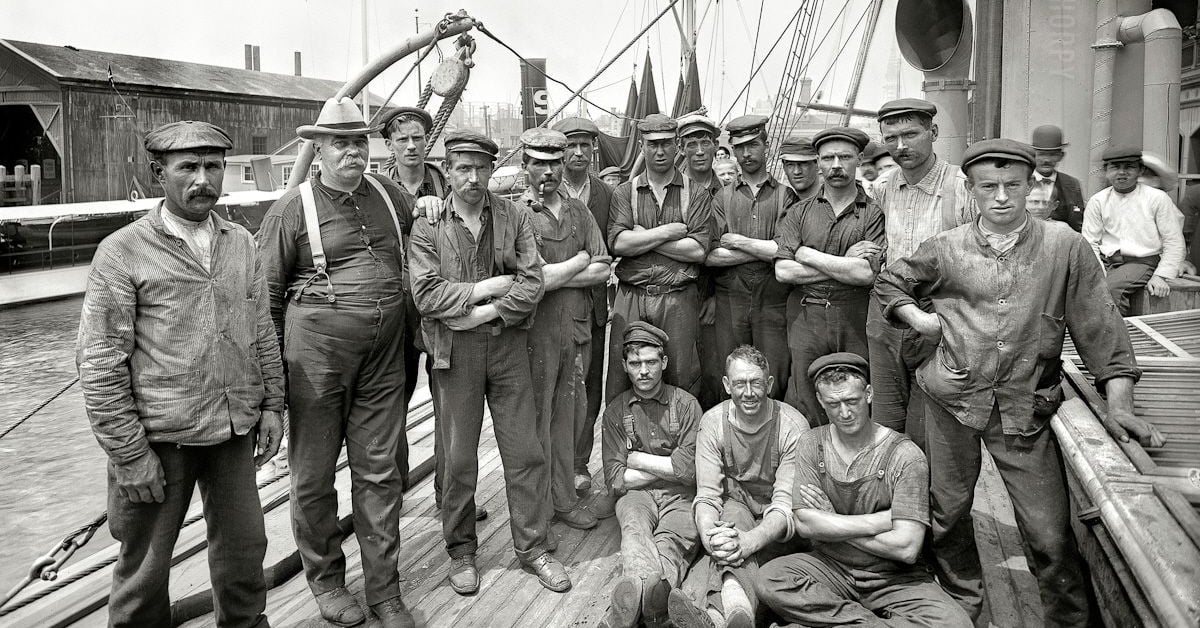A group of men in work clothes and hats pose for a photo on the deck of a boat at a dock. Some stand with arms crossed, while a few sit on the wooden deck. Industrial buildings are visible in the background.