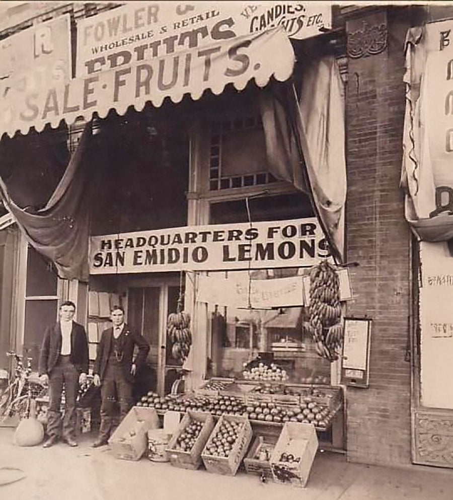 Two men stand outside a vintage storefront with fruit displays and a sign reading "Headquarters for San Emidio Lemons." Bananas and crates of fruit are arranged on the sidewalk; awnings and signs hang above.