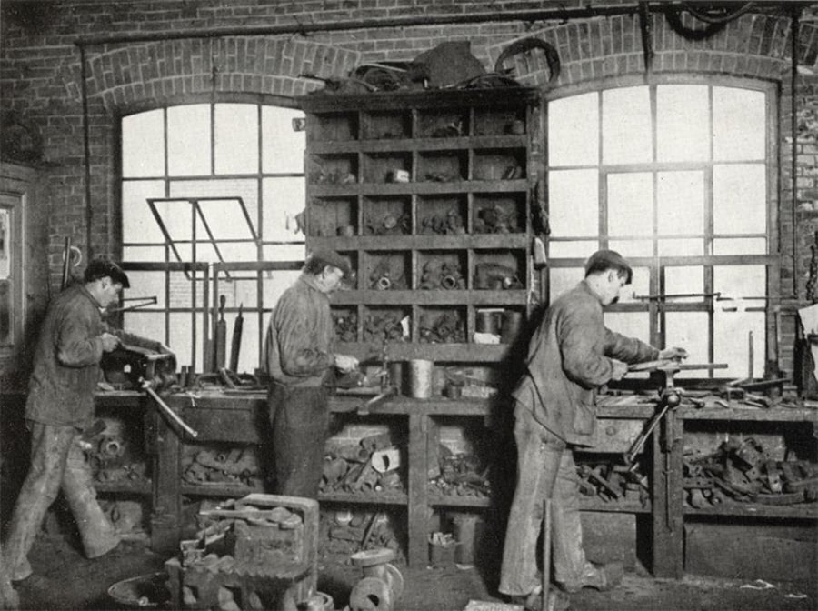 Three men work at a cluttered workbench in a workshop with brick walls and large windows, using tools and surrounded by shelves filled with various mechanical parts.