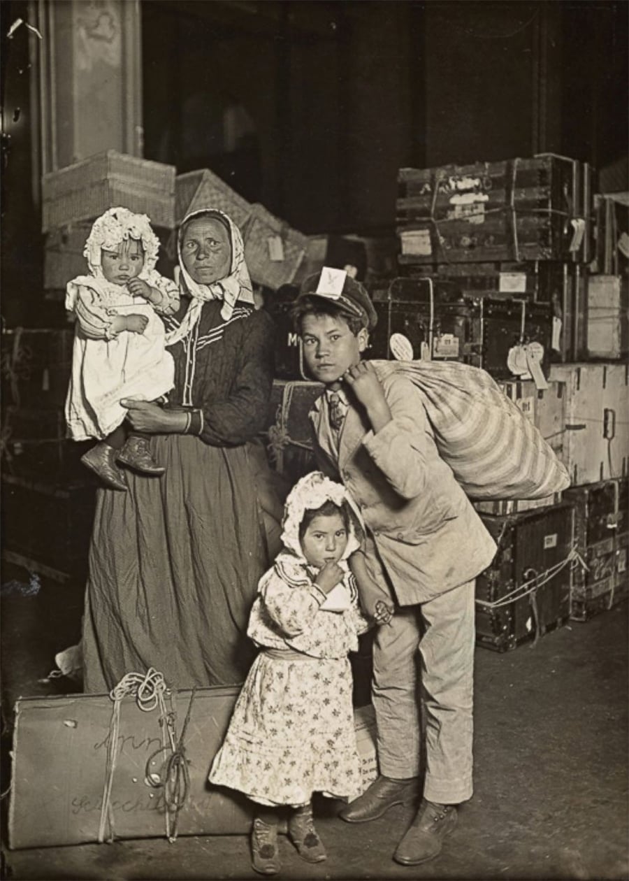 A historical black-and-white photo shows a woman holding a baby, a young man with a sack over his shoulder, and a little girl, all standing in front of stacked luggage, possibly at an immigration station.