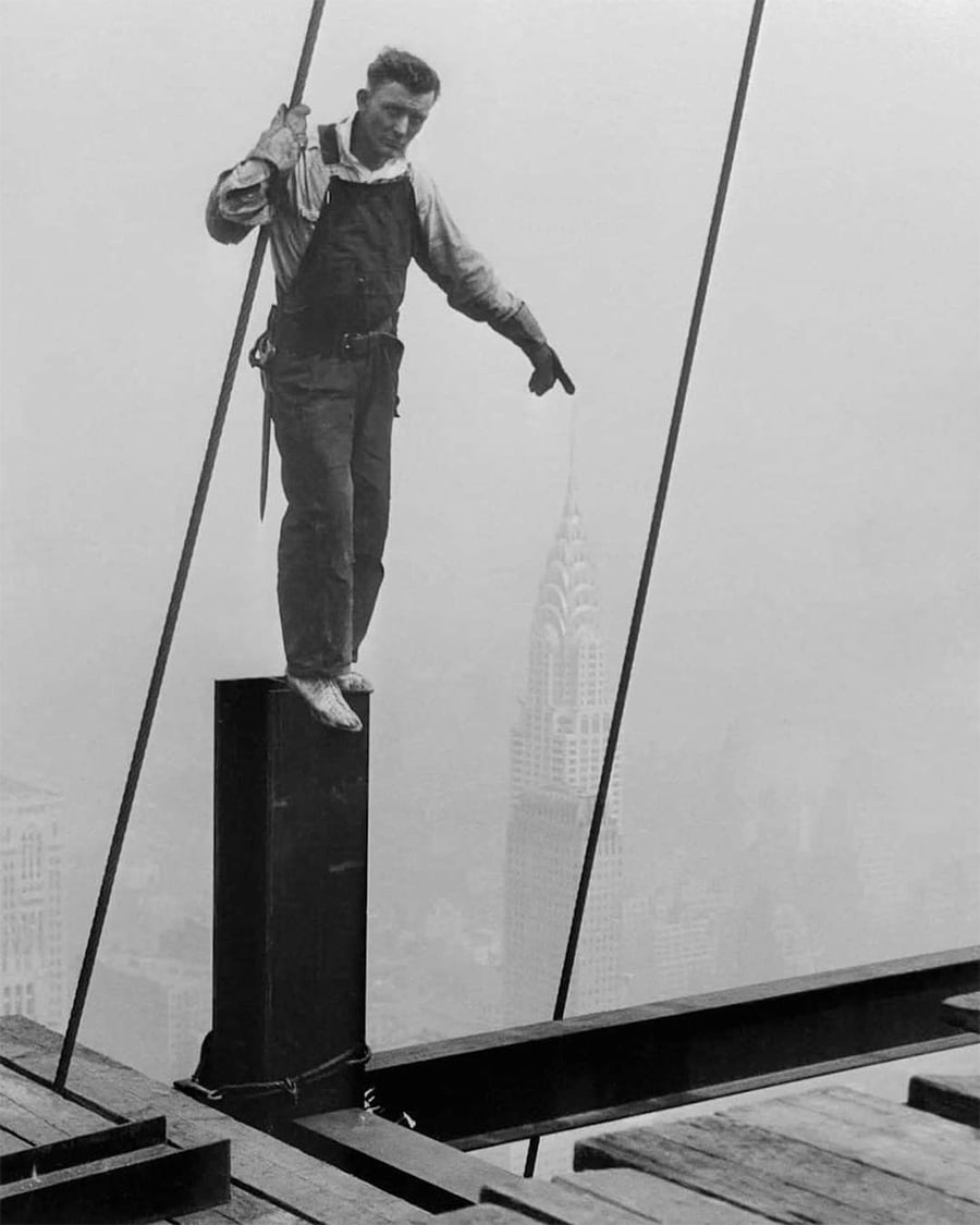 A construction worker balances on a steel beam high above the city with the Chrysler Building visible in the foggy background. He points downward, holding onto a rope for support.