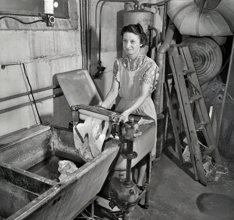 A woman wearing an apron uses a vintage wringer washing machine in a basement laundry room with pipes, a large sink, and utility items in the background.
