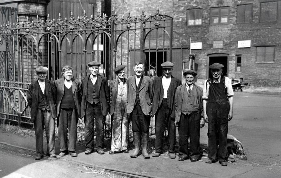 Eight men in work clothes and flat caps stand in a line outside an iron gate, with industrial brick buildings in the background; the scene appears to be from the early 20th century.