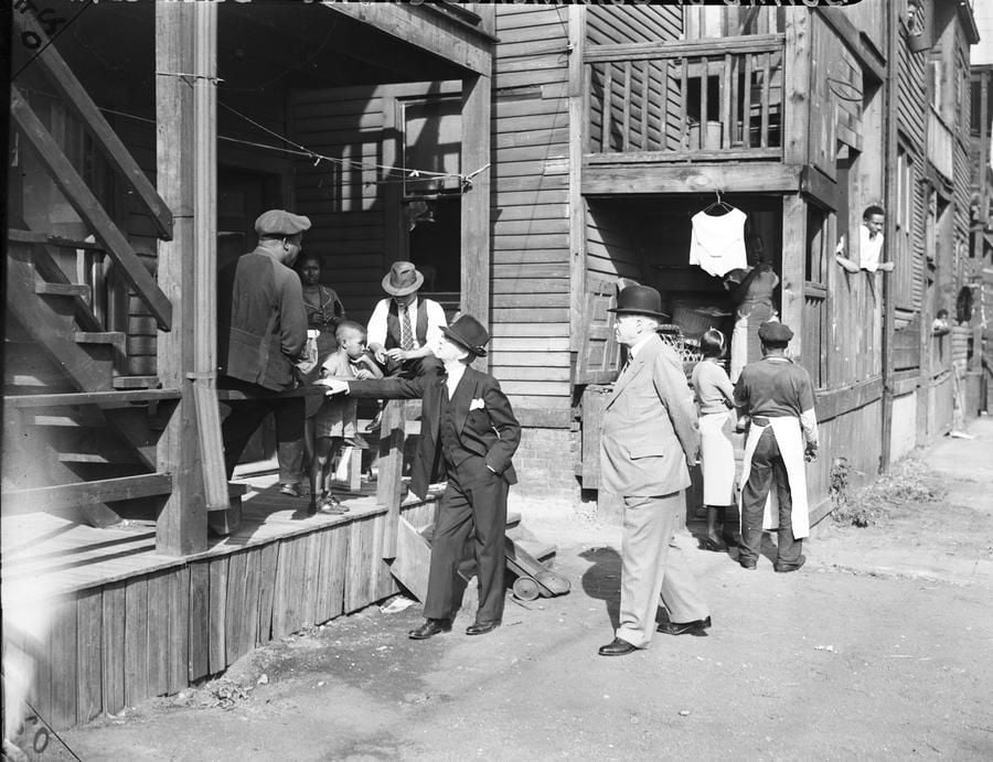 A black-and-white photo shows men in suits and hats talking on a dirt street, while families and children gather outside wooden apartment buildings with balconies, porches, and laundry hanging overhead.