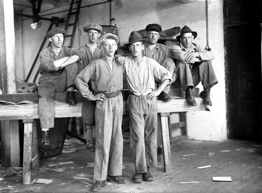 Six men in work clothes and hats pose for a black-and-white photo inside an industrial workshop, with four sitting and two standing in front of a wooden bench. Tools and scattered papers are visible in the background.