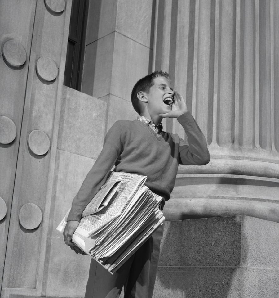 A boy stands outside a large building, holding a stack of newspapers under one arm and shouting, as if calling out headlines. The photo is in black and white.