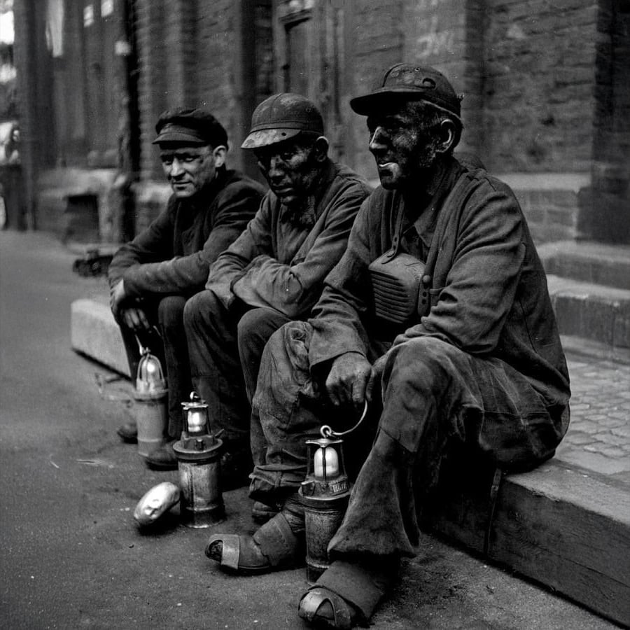 Three coal miners, faces and clothes covered in soot, sit on a curb with their equipment and lanterns, taking a break outside a brick building.