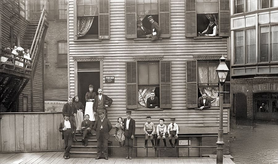 A black-and-white photo of people posing outside and in the windows of a wooden multi-story building in a city. Adults and children stand or sit on stairs, while others lean out of open windows, with city buildings in the background.