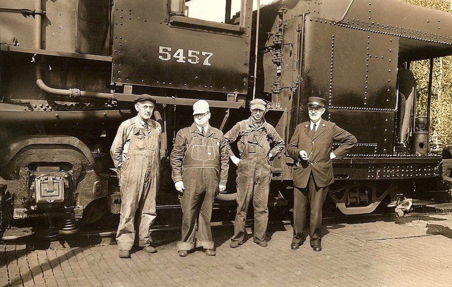 Four men stand in front of a steam locomotive numbered 5457; three wear overalls and work clothes, while one wears a suit and conductor’s hat. The photo appears to be from the early-to-mid 20th century.