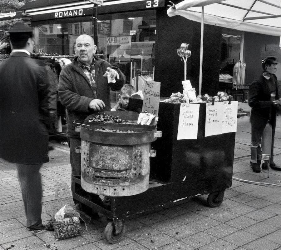 A man stands behind a street cart selling roasted chestnuts, with signs and prices displayed. He gestures while talking to a customer. Another man and a child are nearby, and a shop named "ROMANO" is in the background.