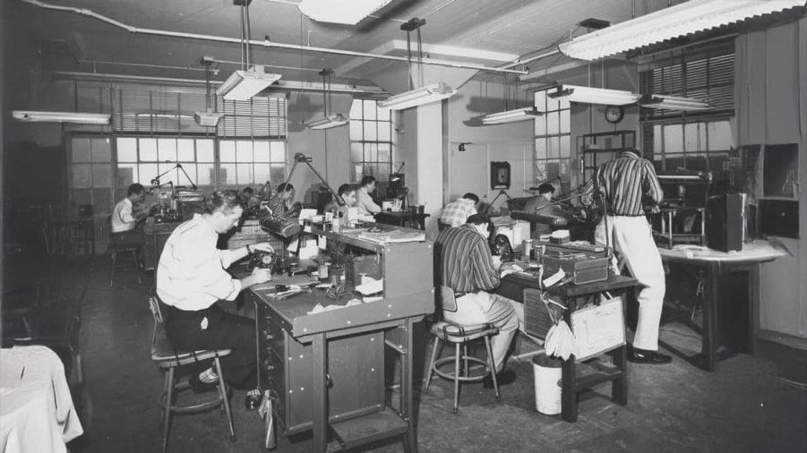 Black and white photo of people working at desks in a factory or workshop, each focused on various tasks. The room is filled with equipment, tools, and overhead lights, with windows lining the back wall.