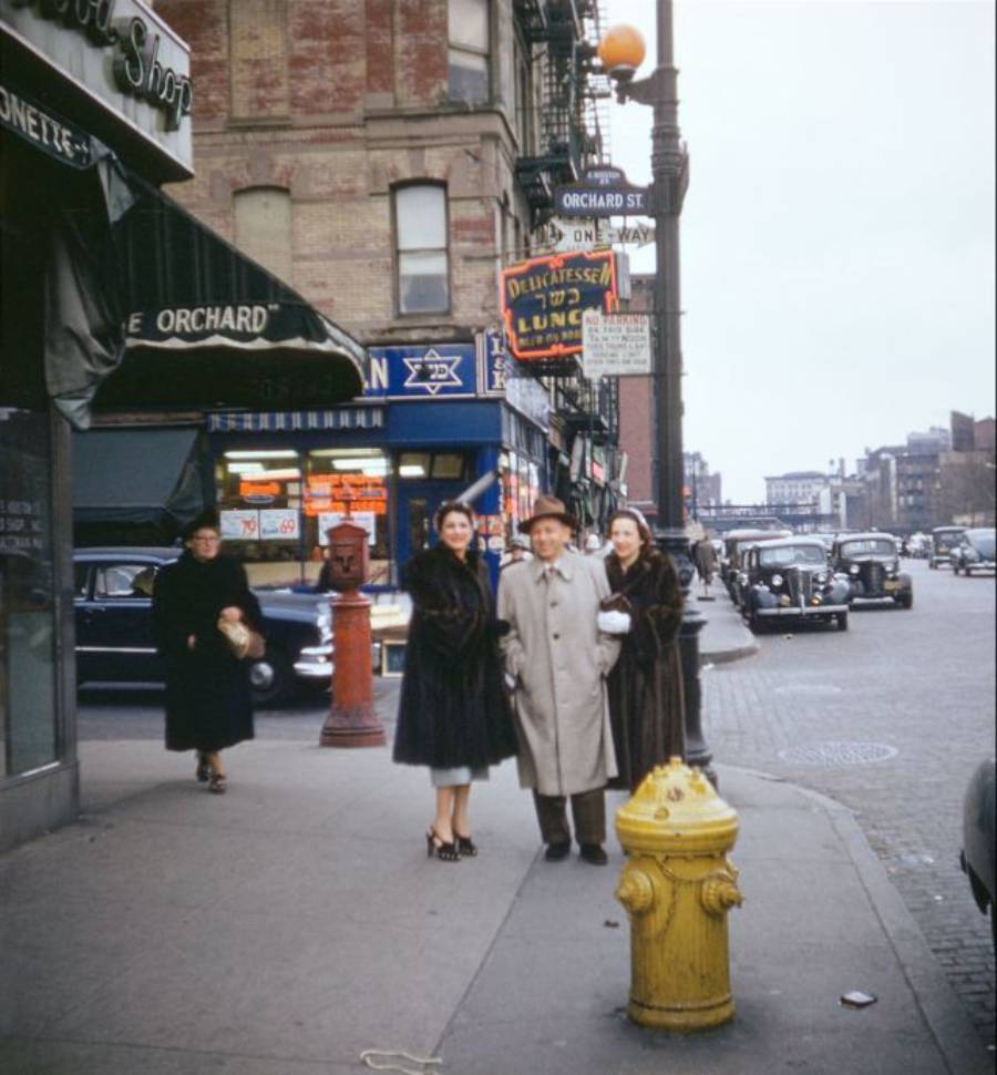 Three people in coats and hats stand on a city sidewalk near a yellow fire hydrant, with storefronts, vintage cars, and street signs visible in the background on a busy urban street.