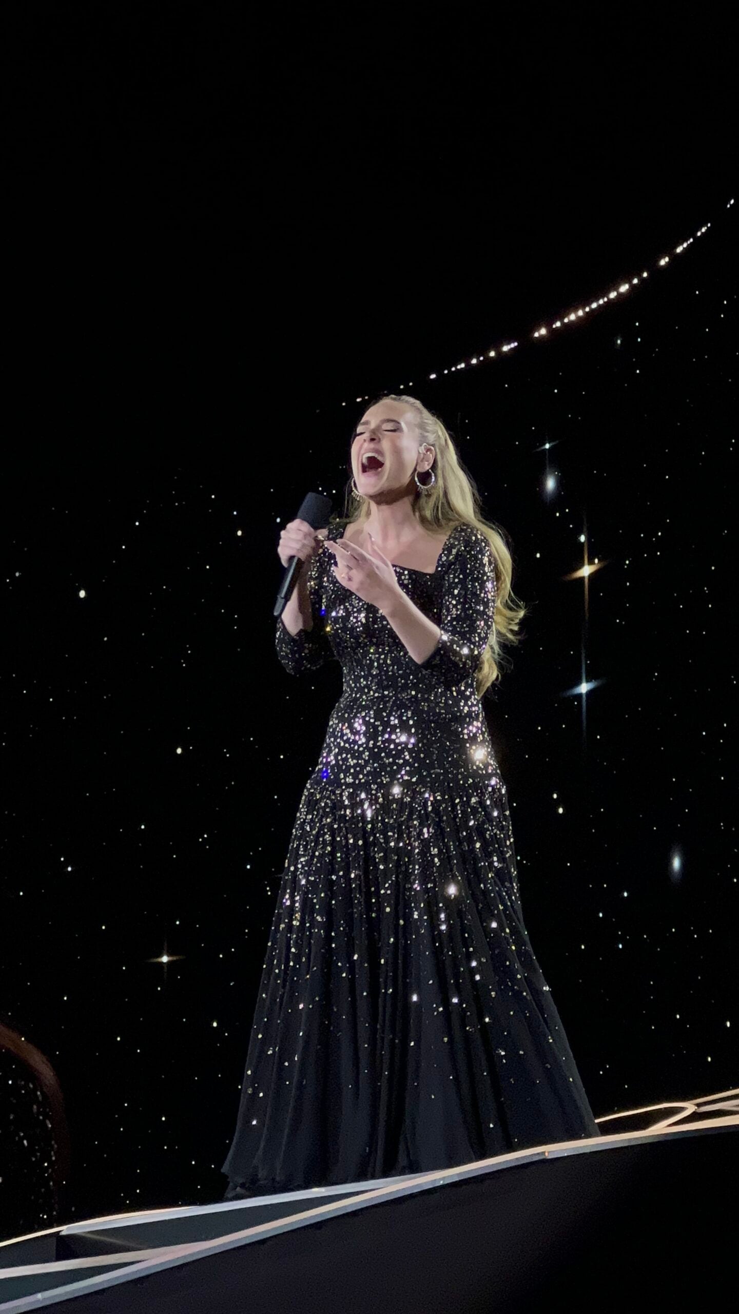 A woman in a sparkling black dress sings passionately on stage, holding a microphone. The background is dark with star-like lights, creating a dramatic, celestial atmosphere.