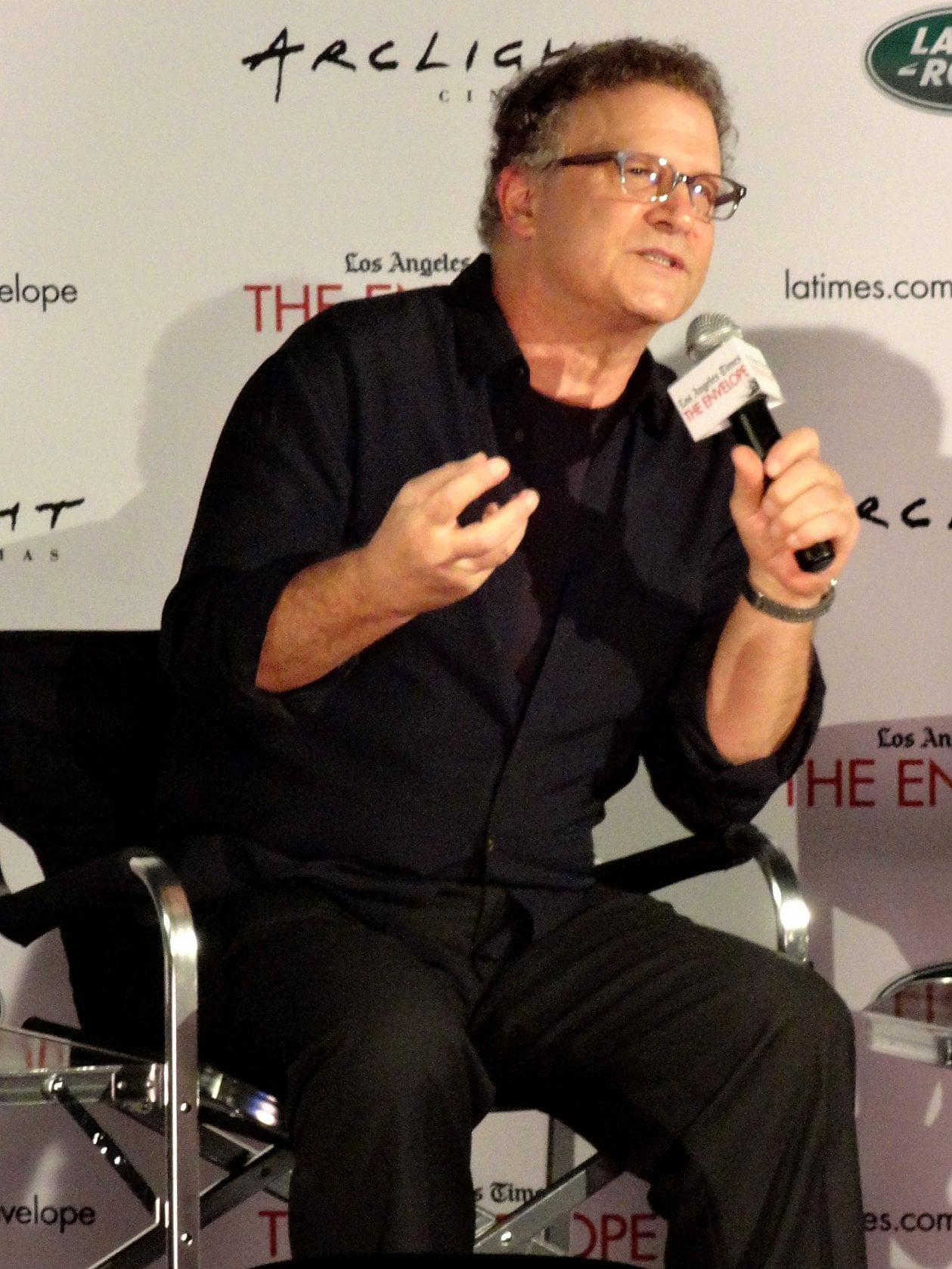 A man wearing glasses and a dark shirt speaks into a microphone while seated on a director's chair at a public event, with a backdrop displaying various logos behind him.