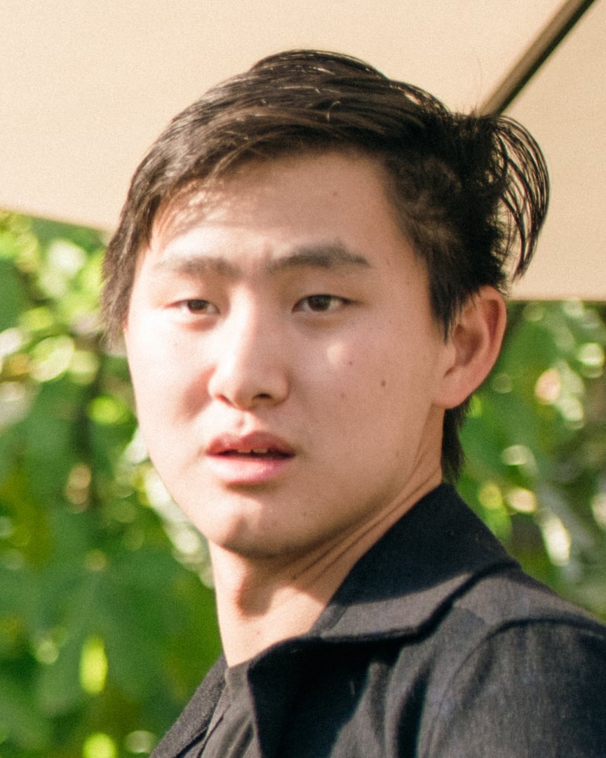 A young person with short dark hair, wearing a black jacket, looks slightly to the side with a neutral expression. Green foliage and a beige umbrella are visible in the blurred background.