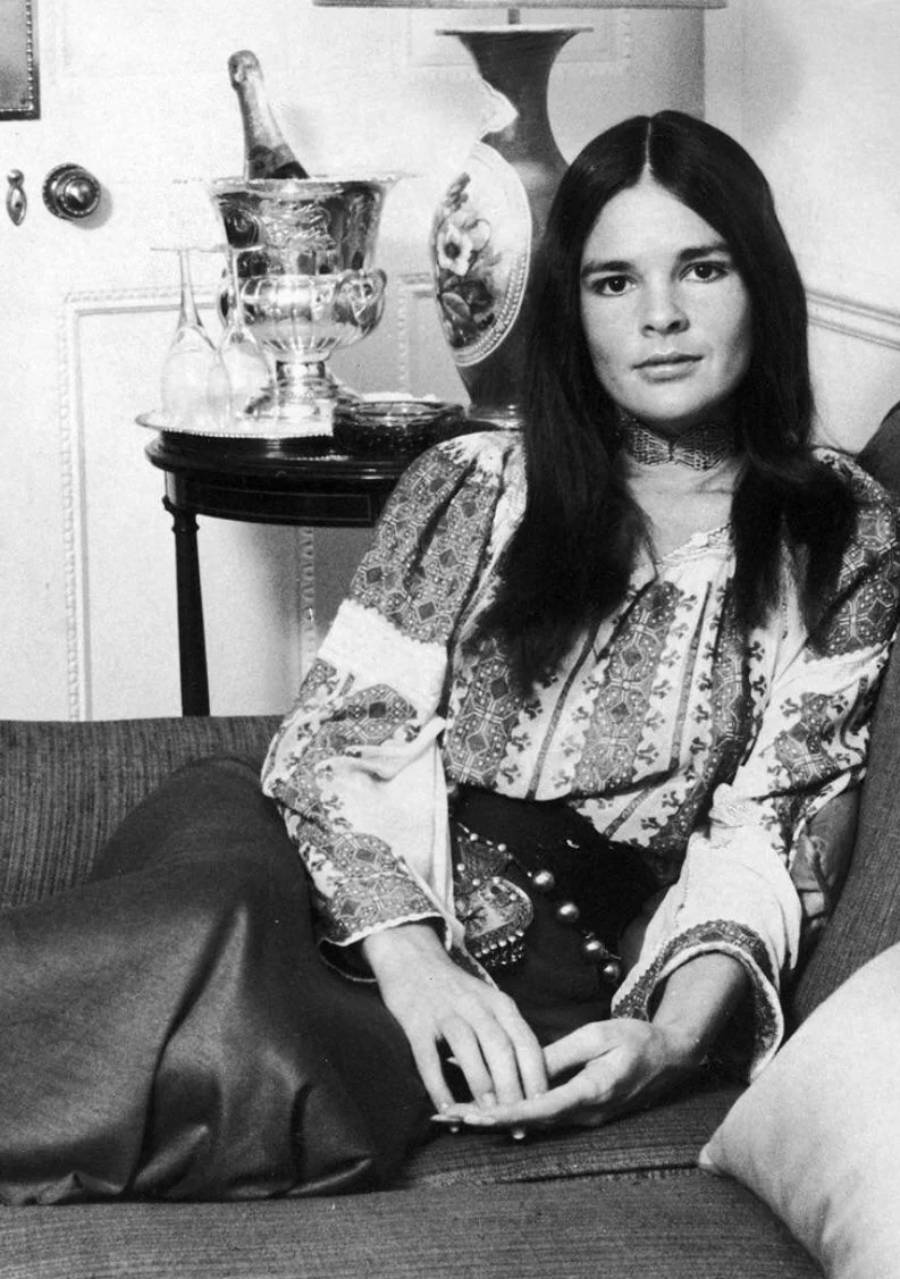 A woman with long dark hair, wearing a patterned blouse and dark skirt, sits relaxed on a sofa. Behind her is a table with a silver ice bucket, glassware, and a lamp. The scene has a vintage, black-and-white look.