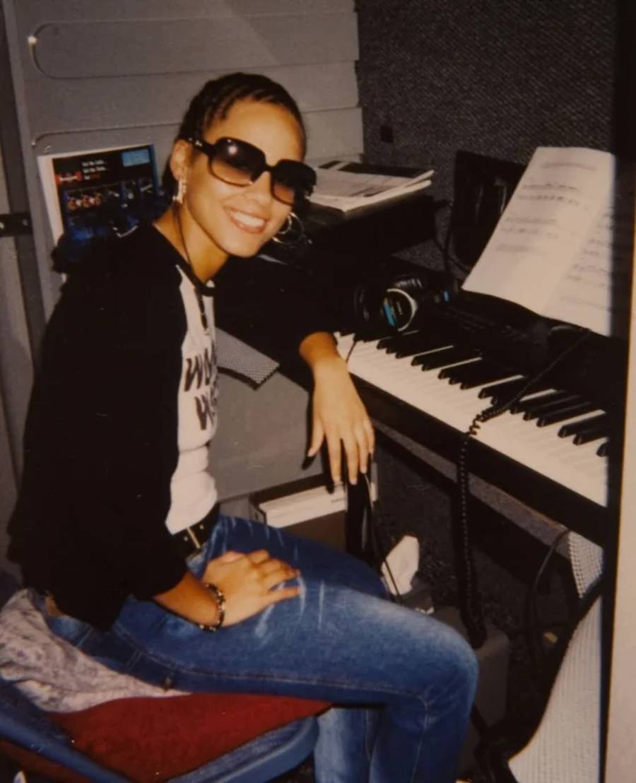 A smiling woman wearing sunglasses and a black-and-white shirt sits at a keyboard in a music studio, with sheet music on the stand and recording equipment nearby.